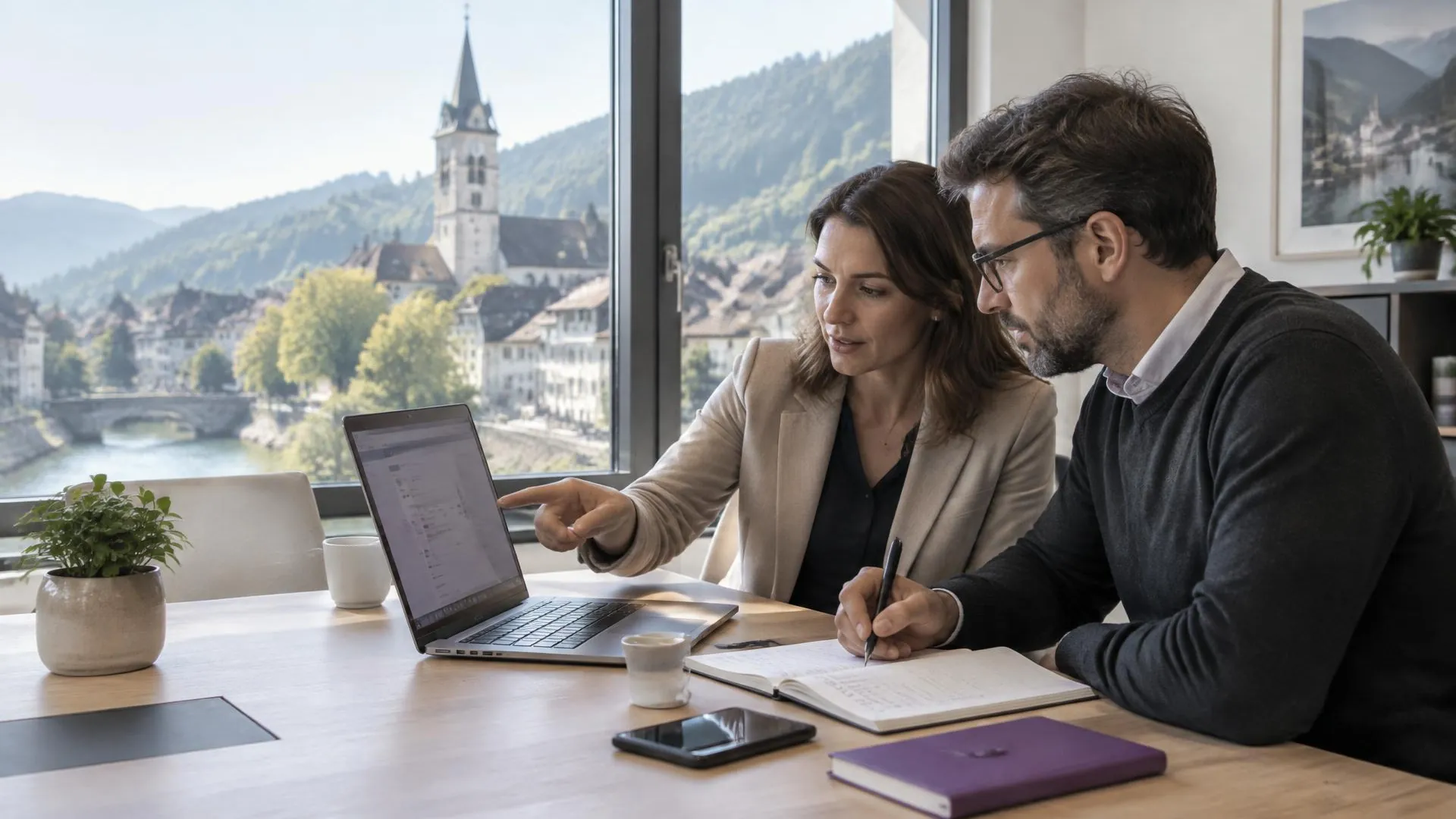 Zwei Fachpersonen planen am Laptop eine Umbenennung und notieren Schritte, im Fenster eine Schweizer Kleinstadt mit Kirchturm am Fluss