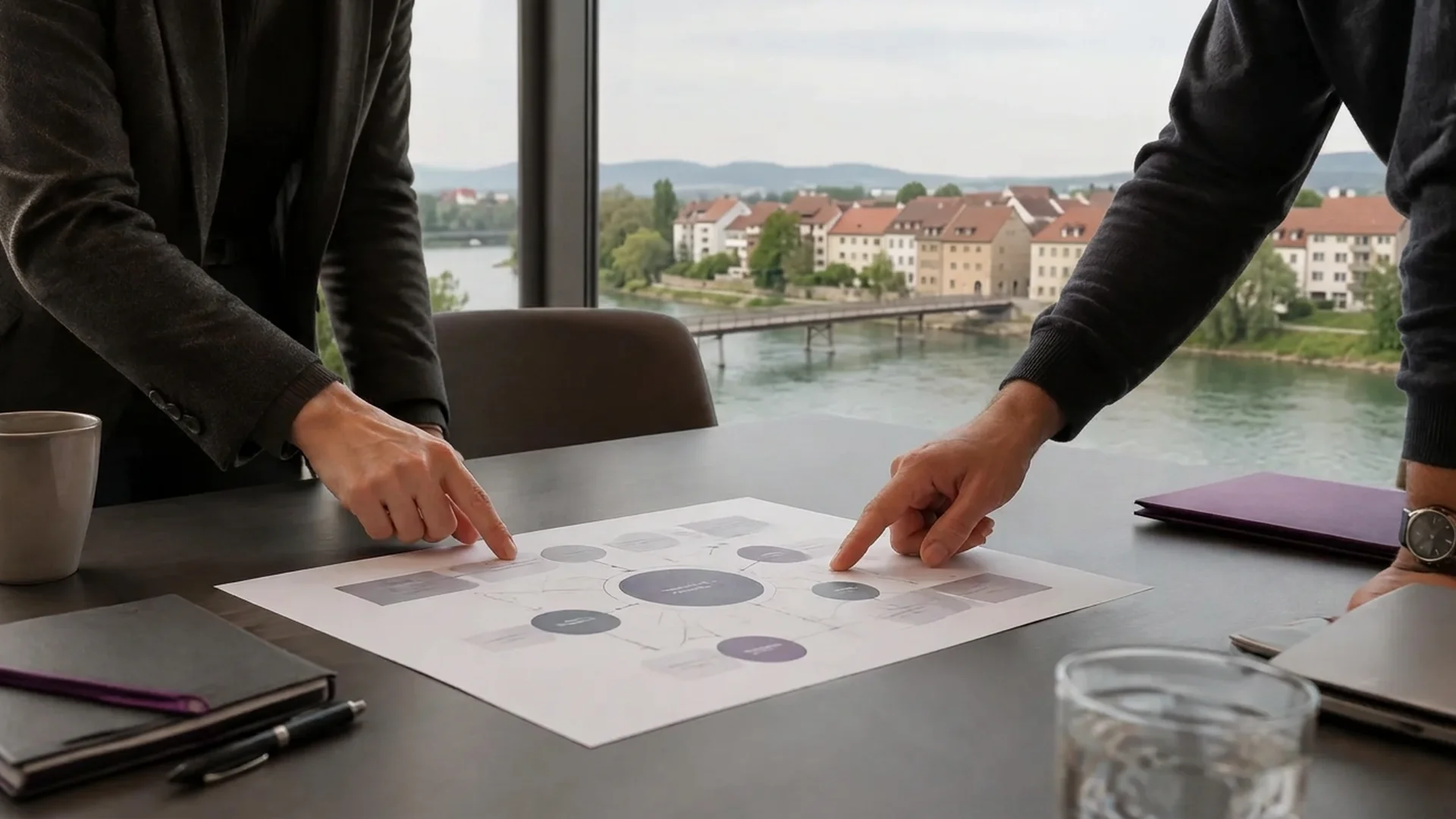 Zwei Personen besprechen ein Schema auf Papier, im Fenster ein Flussufer mit niedrigen Häusern und einer Fussgängerbrücke.