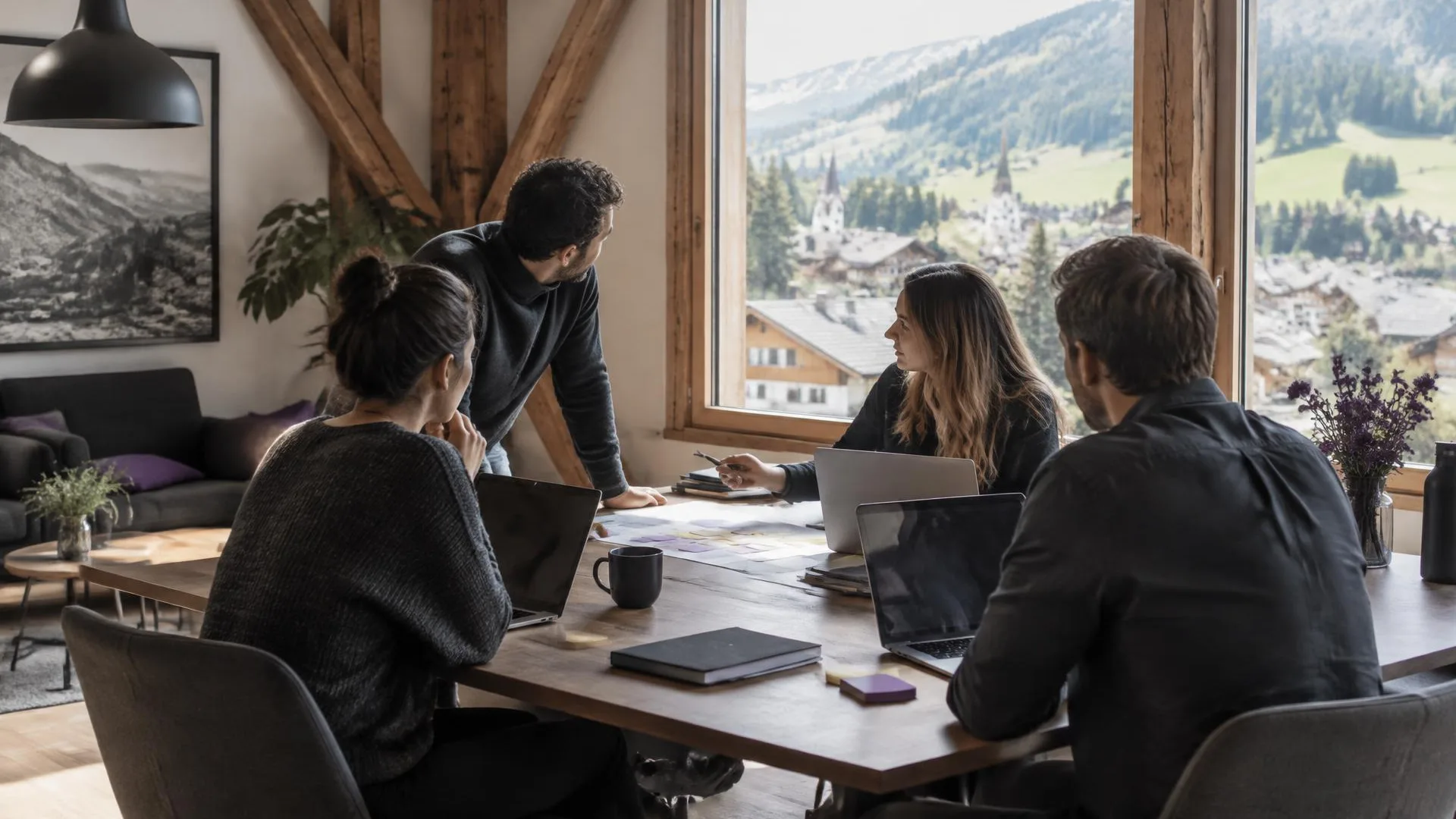 Team arbeitet mit Laptops und Notizzetteln in einem Holzbau, im Fenster ein Schweizer Bergdorf mit Wiesen