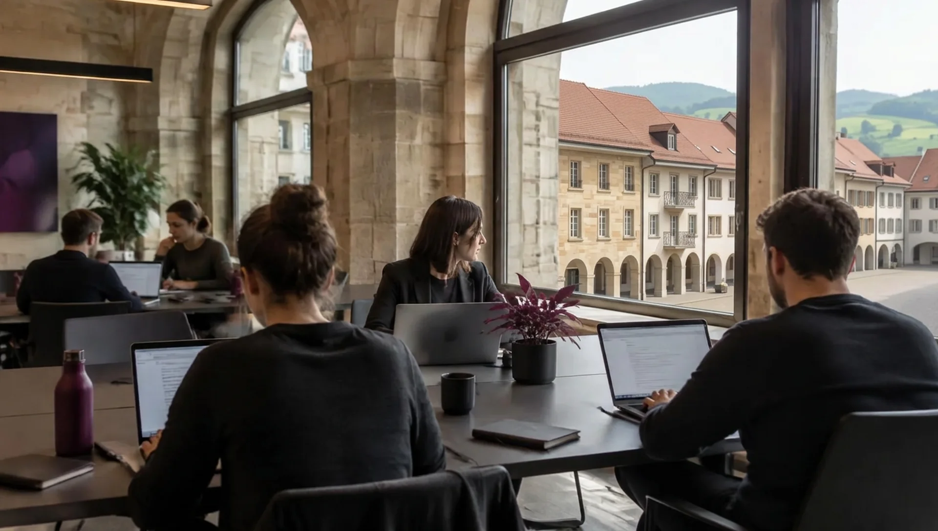 Fünf Personen arbeiten an Laptops in einem Sandsteinraum, im Fenster niedrige Häuser mit Lauben und roten Ziegeldächern.